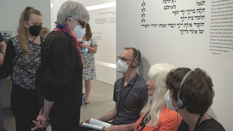 Bei einer Führung durch das Jüdische Museum. Drei Teilnehmende sitzen vor einer Wand mit hebräischen Schriftzeichen. Über Kopfhörer lauschen sie Anja Winter, die vor ihnen steht. Ein Teilnehmer - wie Winter mit FFP2-Maske - hält ein Tastmodell einer Tora-Rolle in den Händen.
